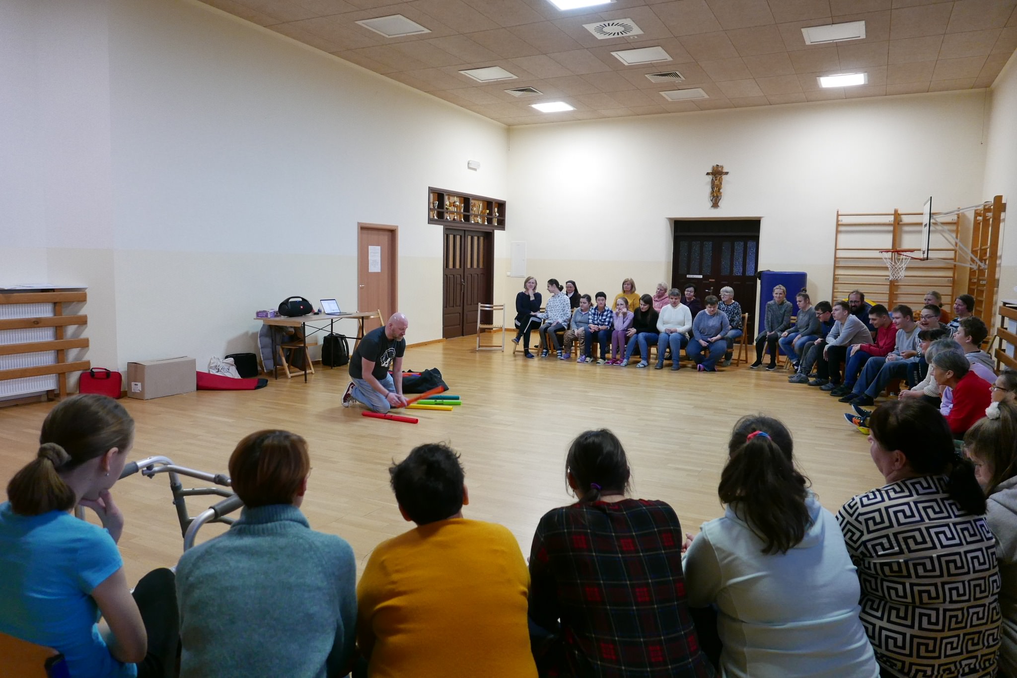 Uczestnicy siedzą w kręgu na sali gimnastycznej z uwagą słuchając pana klęczącego na podłodze i trzymającego w ręce czerwona plastikową rurkę.