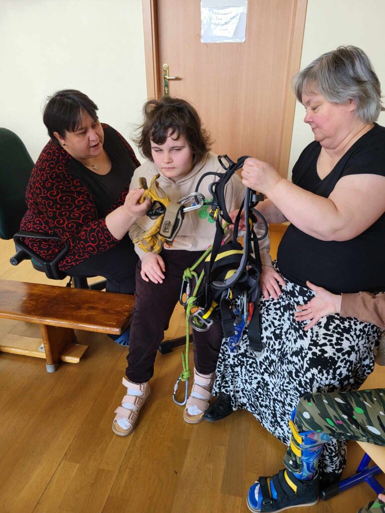 Dziecko siedzi na ławce, a dwie dorosłe osoby pomagają mu założyć uprząż wspinaczkową. Na kolanach trzymają elementy sprzętu wspinaczkowego.