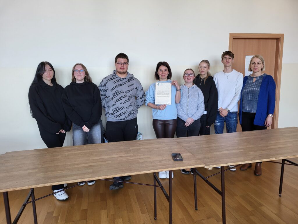 Grupa osób stoi przy drewnianym stole w jasnym pomieszczeniu. Kobieta w środku trzyma dokument, który prezentuje reszcie. Wszyscy patrzą na aparat, a w tle widać drzwi i jasną ścianę.