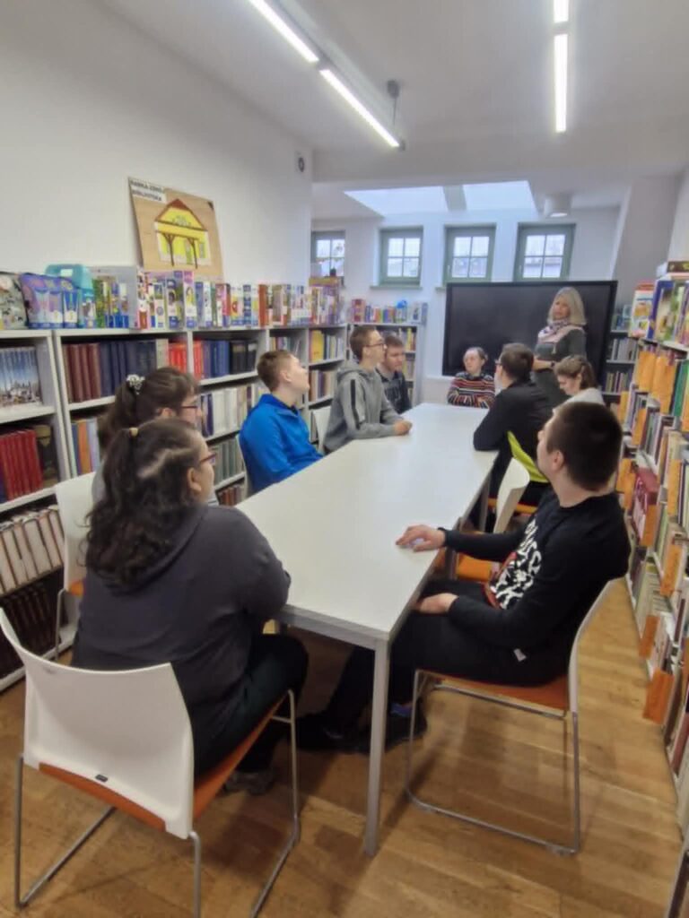 Grupa osób siedzi przy stole uczestnicząc w warsztatach, w tle widać regały z książkami.