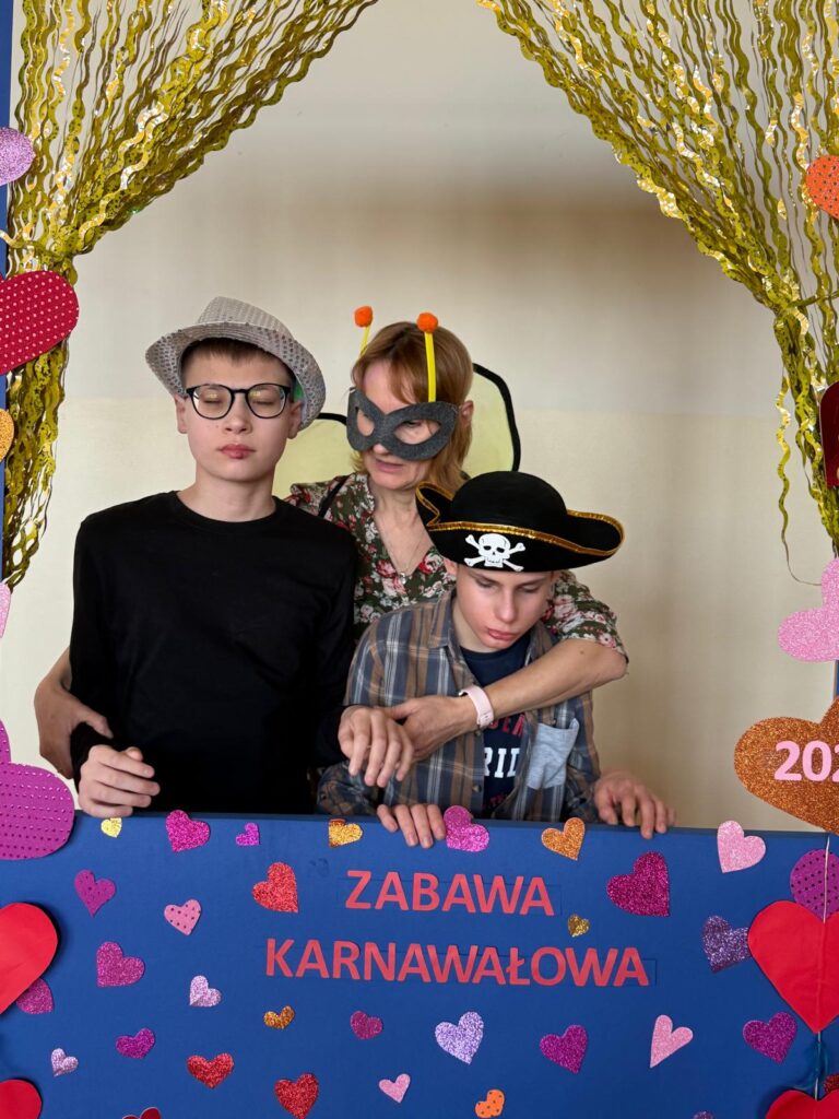 Trzy osoby w karnawałowych przebraniach pozują za ozdobioną ramką z napisem "ZABAWA KARNAWAŁOWA". Tło zdobią złote girlandy i kolorowe serca, tworząc radosną atmosferę.