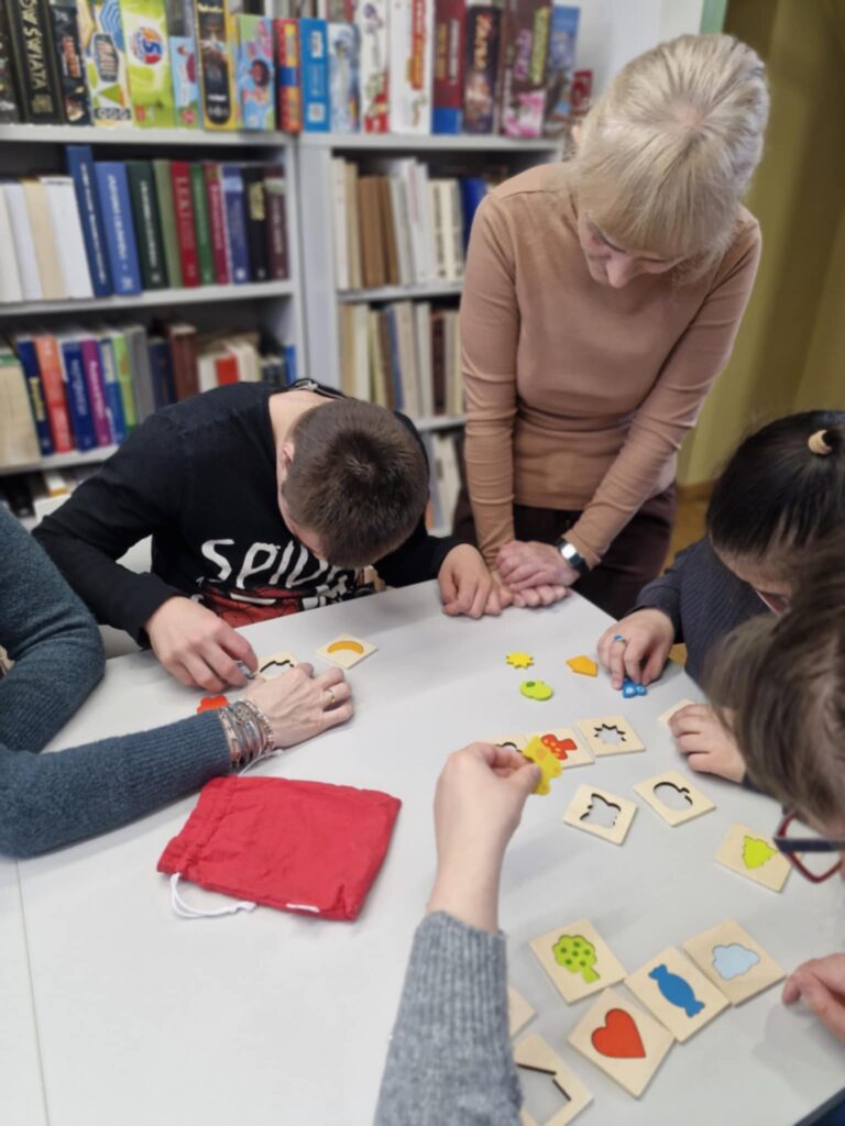 Grupa osób siedzi przy stole i dopasowuje do siebie różne kształty wyciągnięte z czerwonego worka.