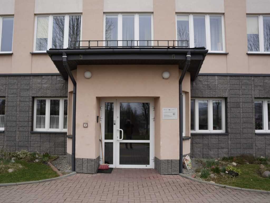 Wejście do budynku z drzwiami szklanymi i daszkiem. Po obu stronach znajdują się okna, a elewacja ma połączenie jasnego tynku i szarych płytek. 