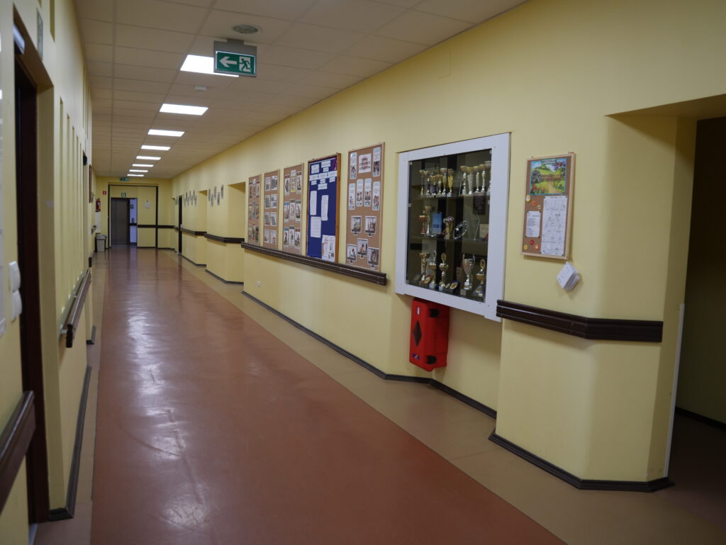 Szkolny korytarz z gablotką z pucharami i tablicami informacyjnymi.