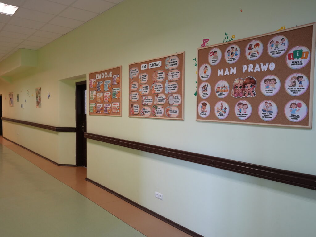 Korytarz szkolny z tablicami edukacyjnymi na ścianie. Tablice zawierają ilustracje i teksty dotyczące emocji,zdrowego odżywiania oraz praw dzieci. Korytarz jest jasny z poręczami wzdłuż ścian.