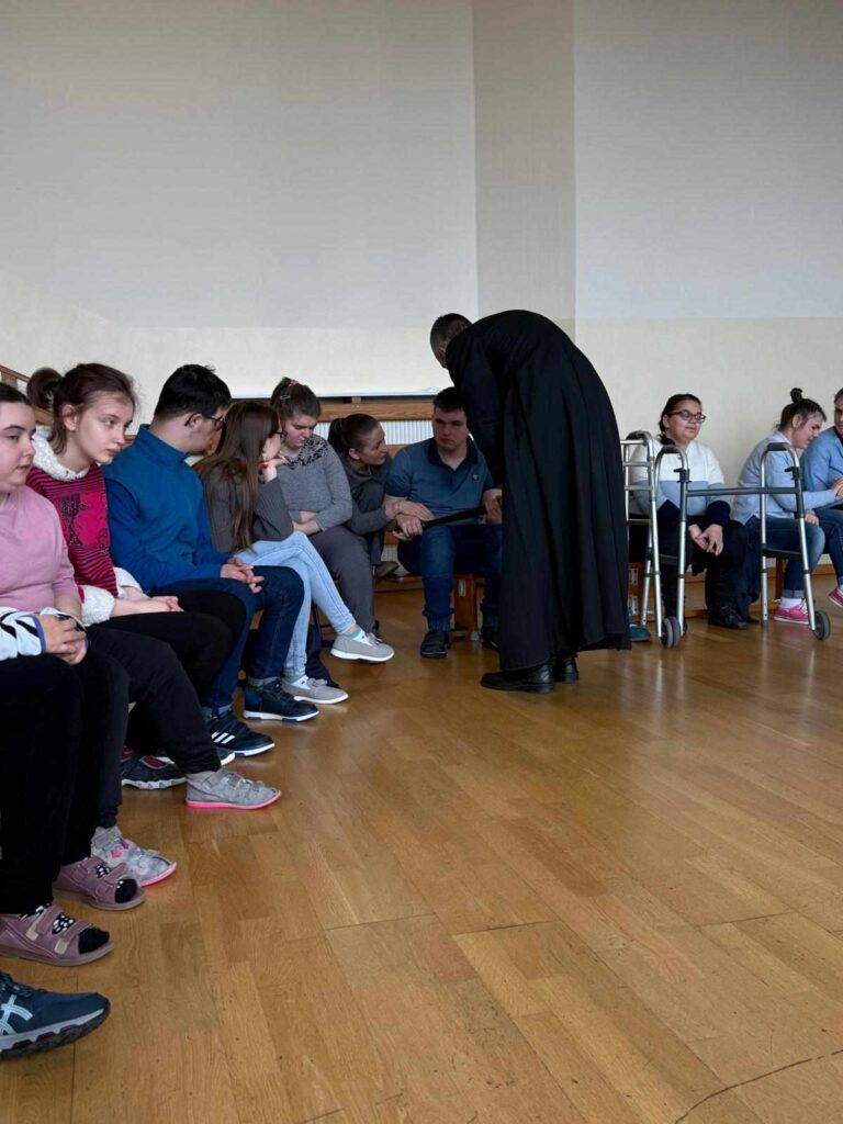 Grupa młodych osób siedzi w półokręgu na ławkach. Wśród nich jest duchowny w czarnej sutannie, pochylony i rozmawiający z jednym z uczestników. 