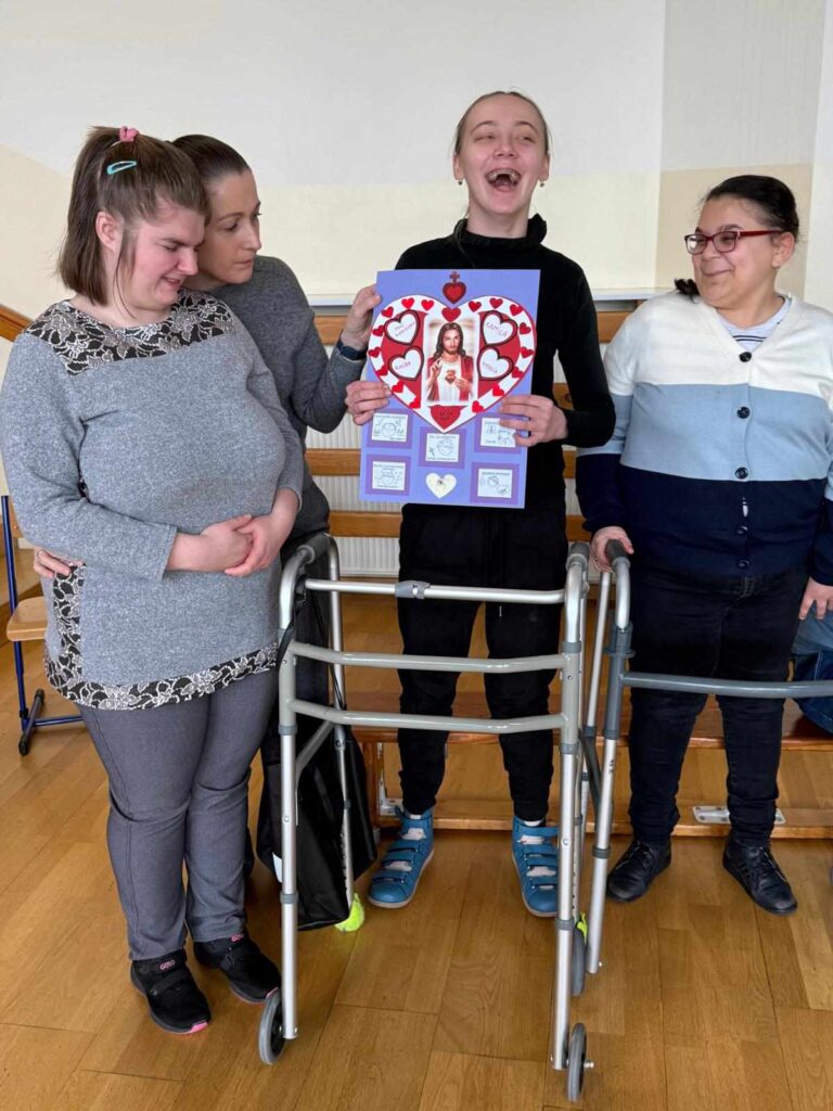 Cztery kobiety stoją razem, z czego dwie korzystają z chodzików. Jedna z nich z uśmiechem trzyma kolorowy plakat z wizerunkiem Jezusa w sercu otoczonym czerwonymi serduszkami i rysunkami.
