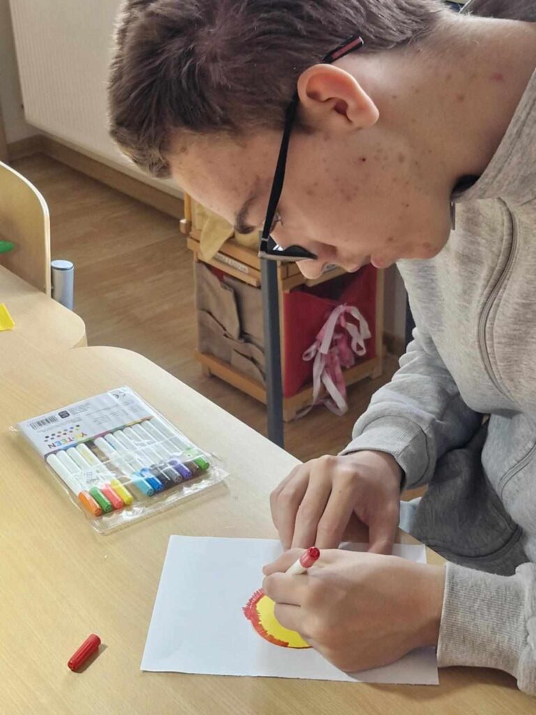 Chłopiec w szarej bluzie i okularach, maluje obrzeże żółtego koła czerwonym pisakiem siedząc przy stoliku.