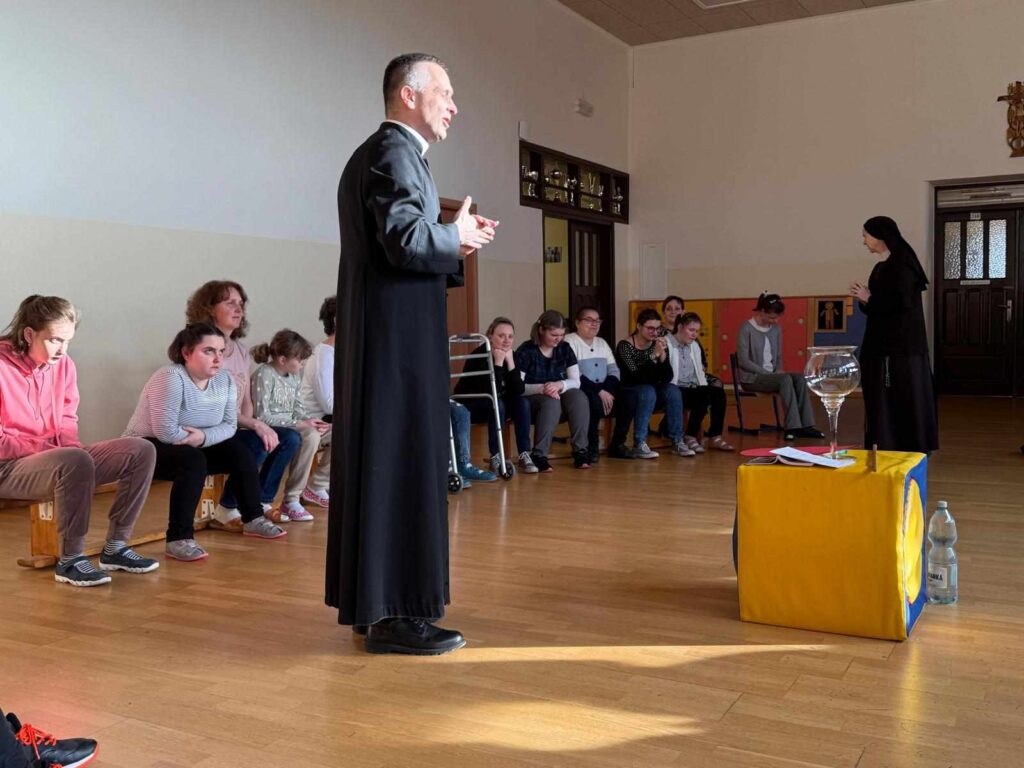 Kapłan w czarnej sutannie stoi na środku sali. Wokół niego siedzi grupa osób, uważnie go słuchając.