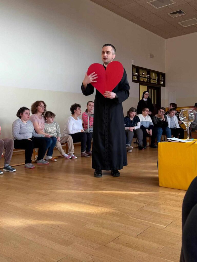 Ksiądz stoi na środku sali i trzyma duże czerwone serce. wokół niego siedzi grupa dzieci i dorosłych, uważnie go słuchając.