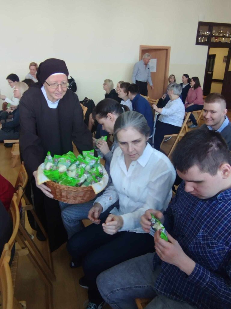 Siostra Zakonna rozdaje uczestnikom spotkania słodycze z koszyka. Ludzie siedzą na krzesłach w sali i z uśmiechem przyjmują poczęstunek.