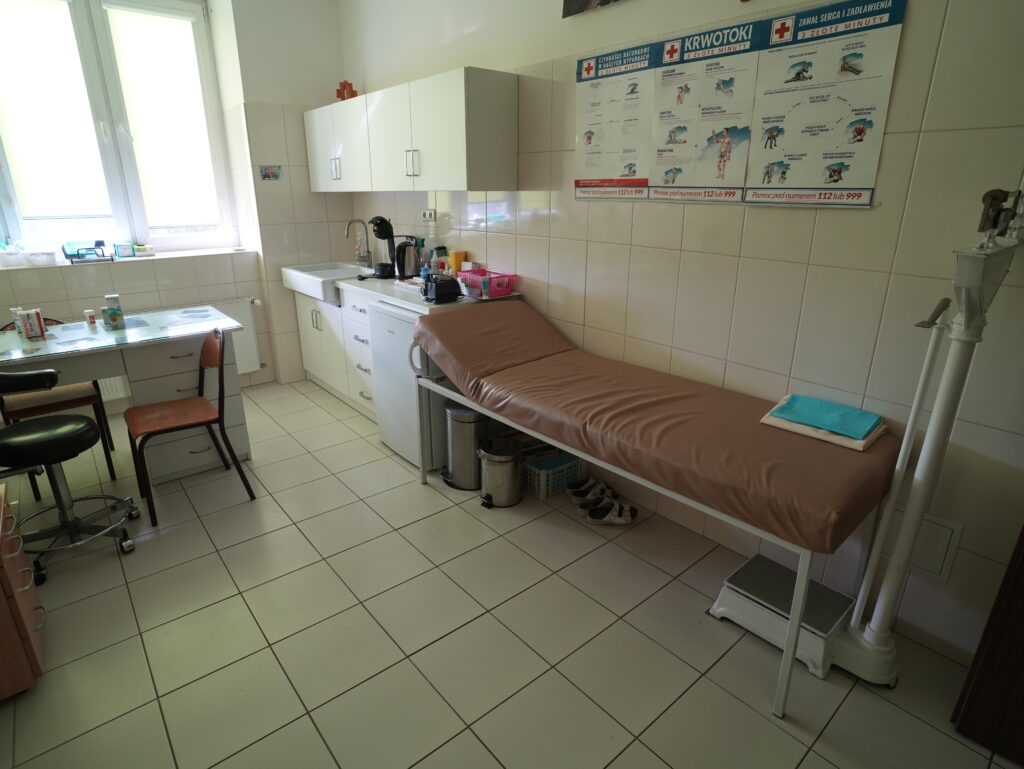 Gabinet medyczny z kozetką, wagą, szafkami i biurkiem.