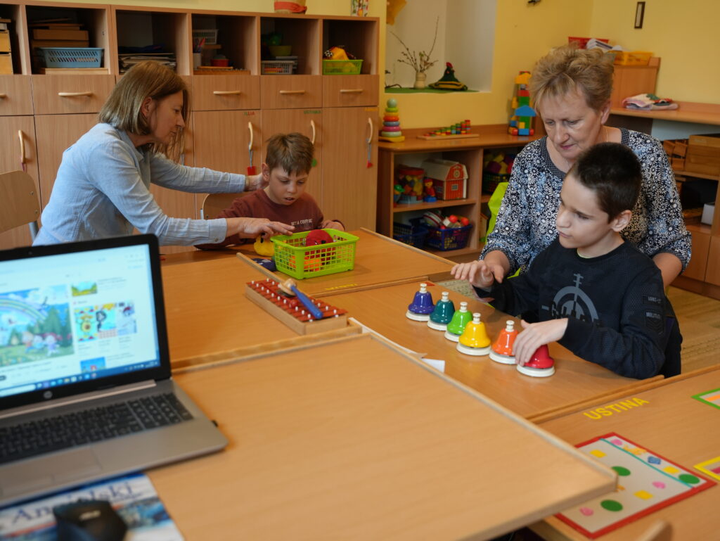 Dwoje dzieci z nauczycielkami siedzą przy stolikach w klasie. Chłopiec w niebieskiej bluzie gra na dzwonkach naciskowych, drugi chłopiec wyciąga rzeczy z koszyka.