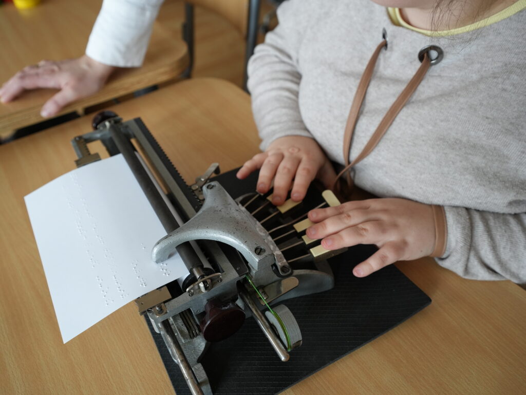Dziewczynka przy stoliku w klasie pisze na maszynie brajlowskiej.