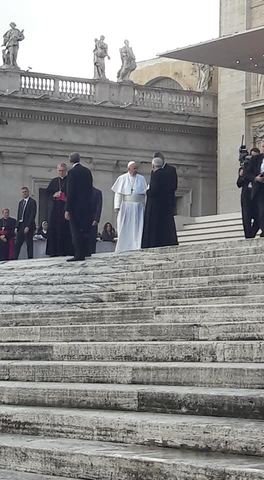 Grupa duchownych, w tym papież Franciszek, stoi na marmurowych schodach przed monumentalnym budynkiem z rzeźbami na balustradzie. 