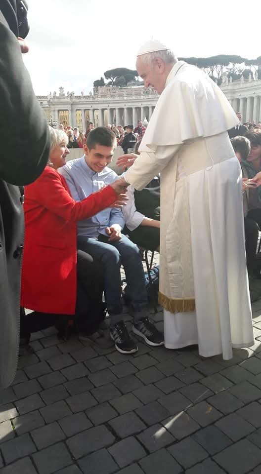 Papież Franciszek pochyla się, ściskając ręce  kobiety w czerwonym płaszczu i młodego mężczyzny, wśród zgromadzonych ludzi na placu przed kolumnadą watykańską.