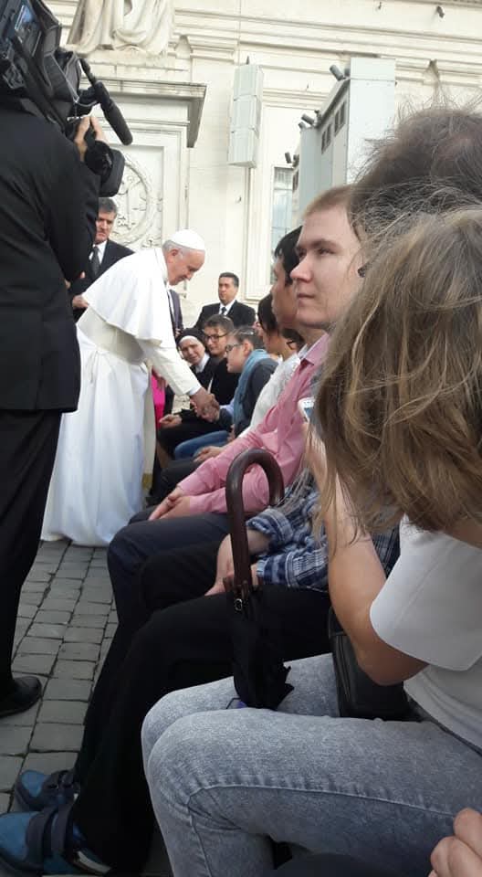 Papież Franciszek pochyla się, aby uścisnąć rękę jednej z wiernych siedzących w rzędzie na kamiennym placyku, obok stoi kamerzysta i ochroniarze, w tle widać fasadę watykańskiego budynku.