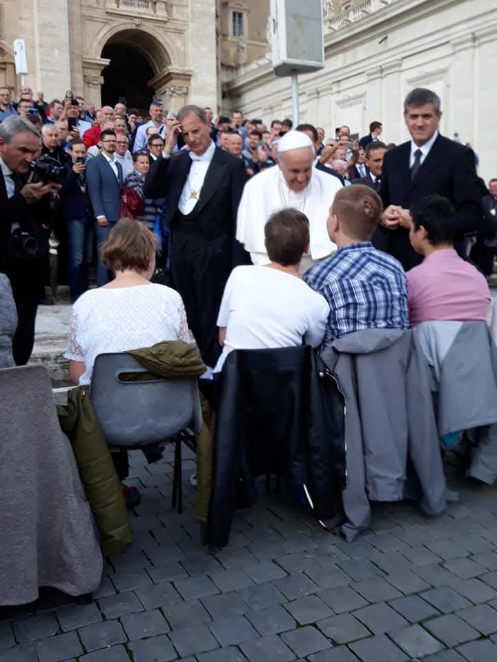 Papież wita się z grupą młodych ludzi siedzących na krzesłach podczas spotkania na świeżym powietrzu. W tle stoi tłum ludzi i ochrona.