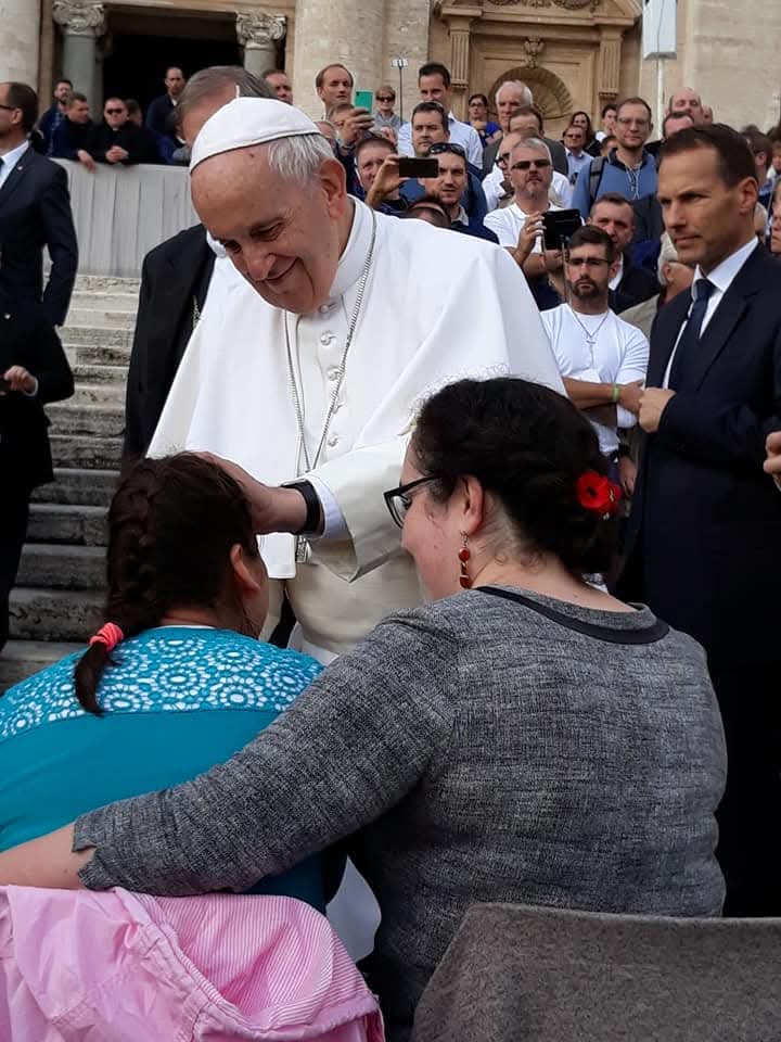 Papież uśmiecha się i błogosławi dziewczynkę kładąc jej rękę na głowie, obok siedzi kobieta. W tle zgromadzony tłum.
