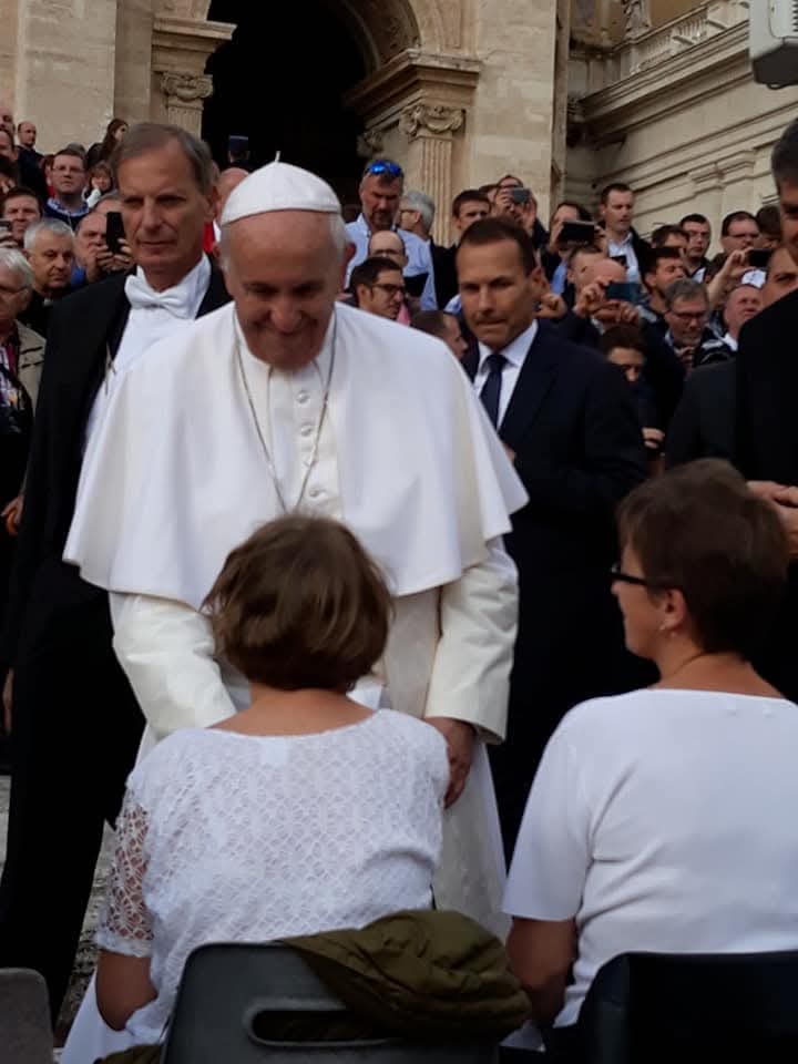 Papież rozmawia z dwiema osobami siedzącymi przed nim, w tle tłum ludzi i ochrona.