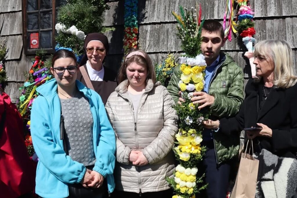 Grupa osób stoi przed drewnianym budynkiem z kolorowymi palmami wielkanocnymi; jedna osoba trzyma dużą palmę z żółtymi i białymi kwiatami.