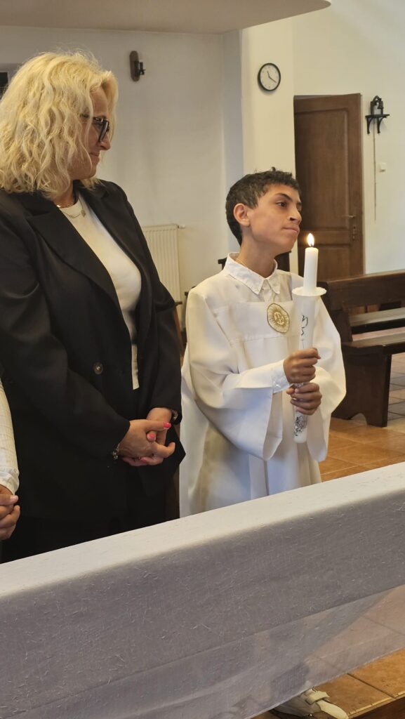 Chłopiec w białej szacie liturgicznej trzyma zapaloną świecę podczas ceremonii Pierwszej Komunii Świętej. Obok niego stoi kobieta w czarnej marynarce i białej bluzce, patrząc na niego z dumą. W tle widać ławki, zegar ścienny i drzwi.