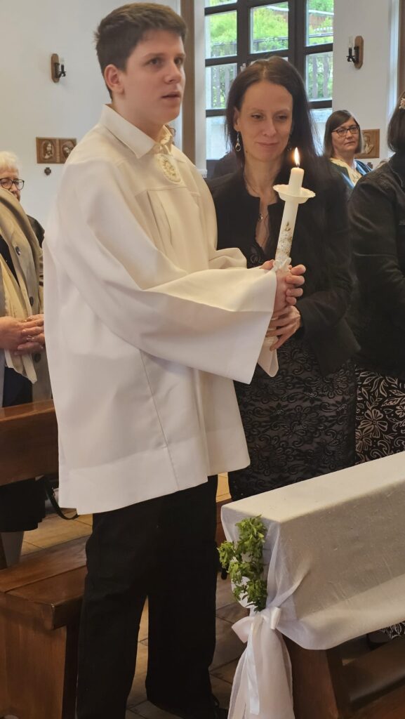 Chłopiec w krótkiej albie komunijnej trzyma zapaloną świecę obok niego stoi kobieta i pomaga mu w trzymaniu świecy.