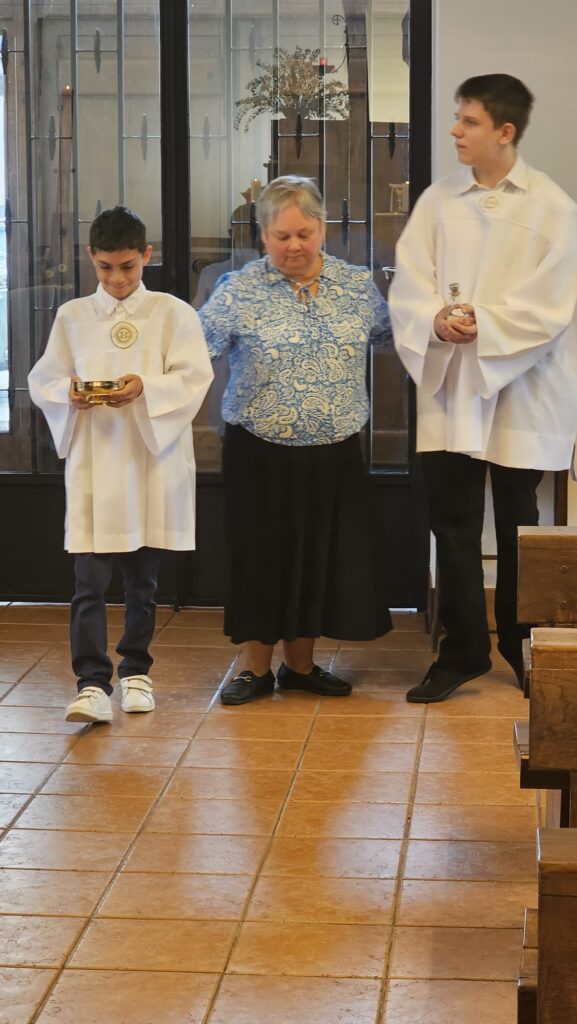 Dwaj chłopcy w albach i kobieta przygotowują dary ofiarne podczas Mszy Świętej.