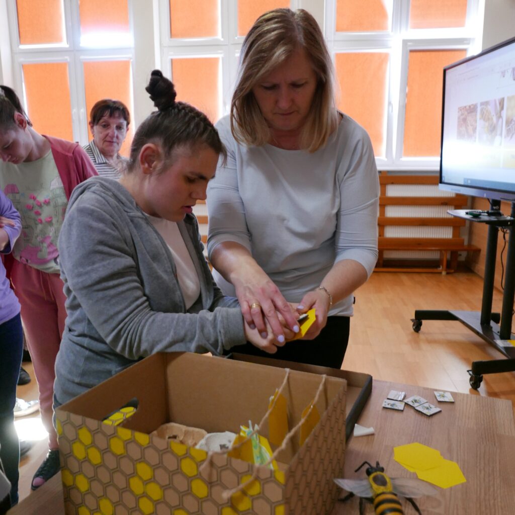 Młoda kobieta z pomocą nauczycielki nakleja obrazki na papier. Na stole leżą kartki , obrazki, model pszczoły i ula.