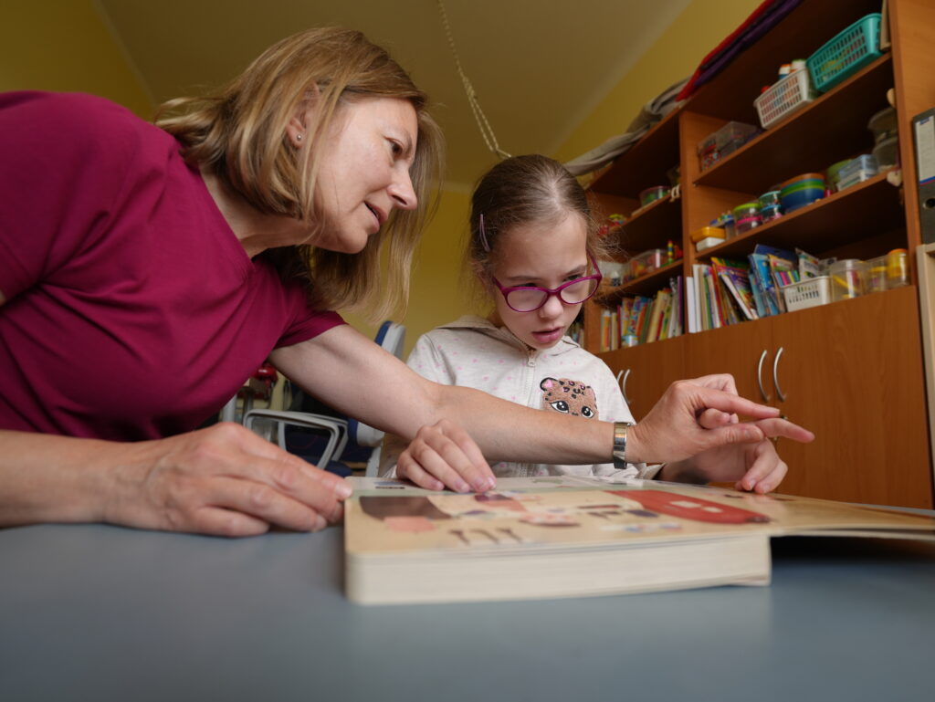Nauczycielka pracuje indywidualnie z uczennicą, wspólnie przeglądają książkę obrazkową. Dziewczynka uważnie słucha i śledzi ilustracje. W tle widać regały z pomocami edukacyjnymi – scena pełna ciepła i zaangażowania.