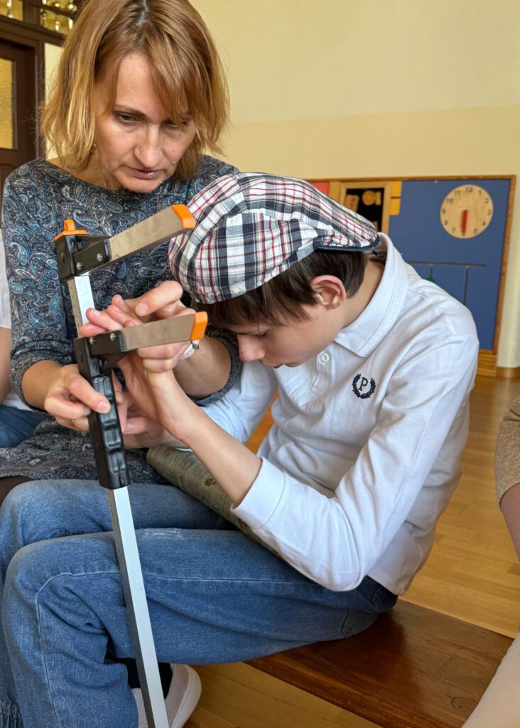 Chłopiec w czapce ogląda przyrząd do mierzenia grubości drzewa, wspierany przez kobietę trzymającą jego dłonie.