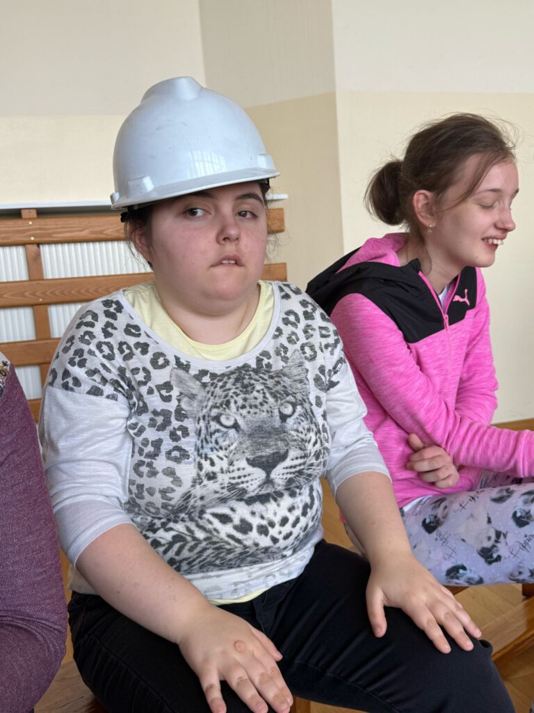 Dwie dziewczyny siedzą na ławce; jedna ma na głowie biały kask i bluzkę z lampartem, druga się uśmiecha.