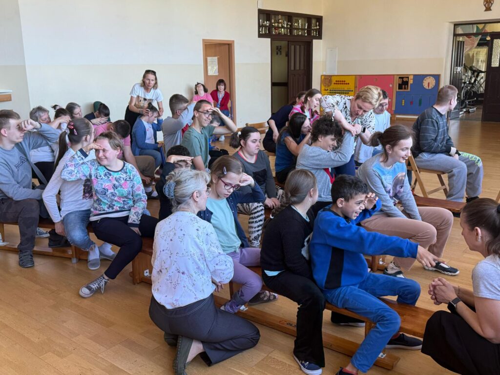 Grupa osób siedzi na ławkach jedno za drugim wykonując polecenia prowadzących.