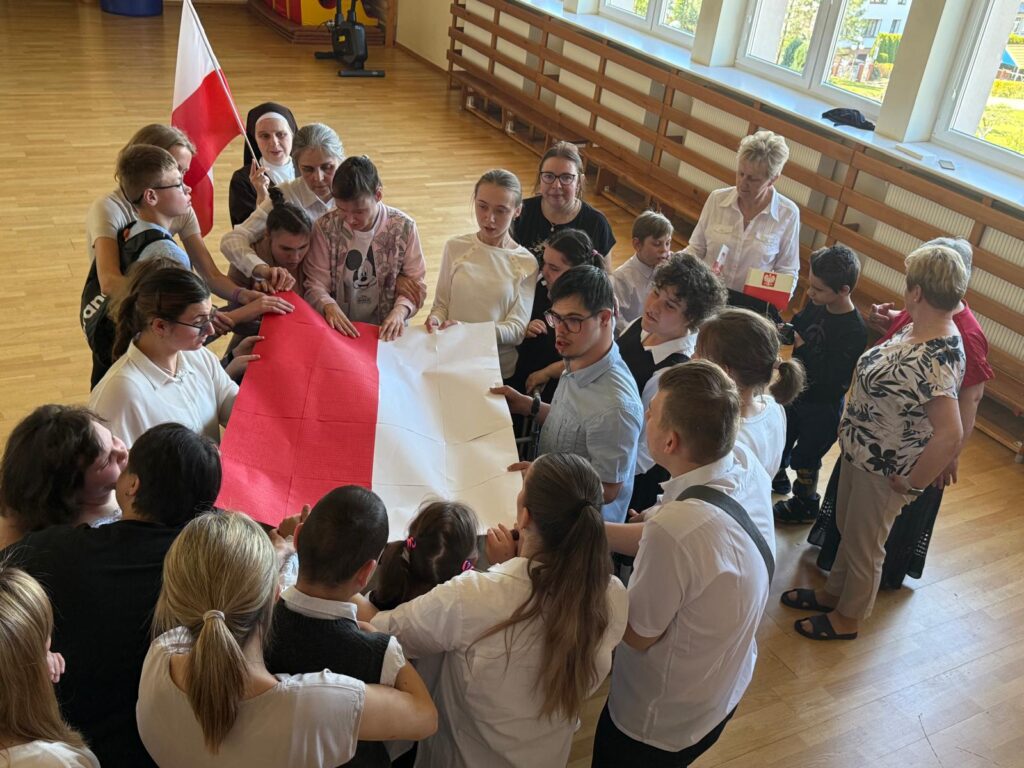 Grupa młodzieży i dorosłych trzyma w rękach dużą biało-czerwoną flagę z papieru.
