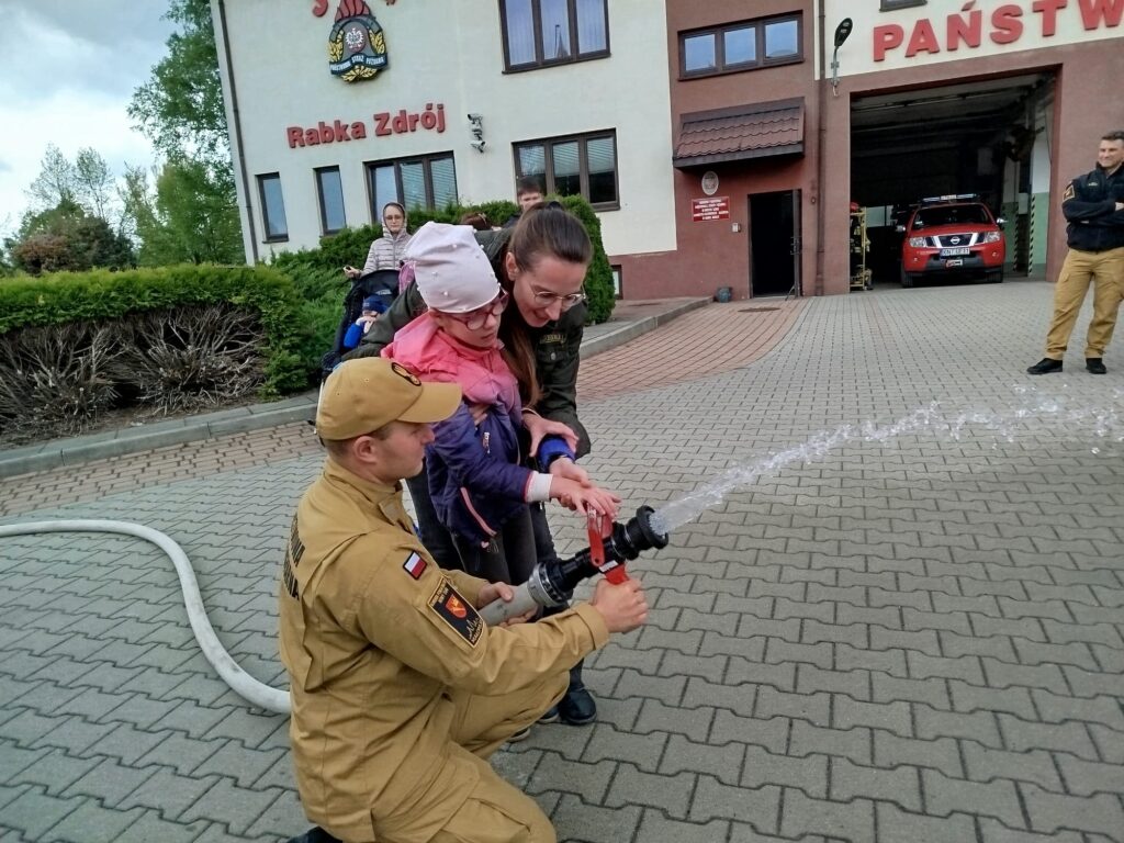 Dziewczynka z nauczycielką i strażakiem w mundurze leją wodę z węża strażackiego. 