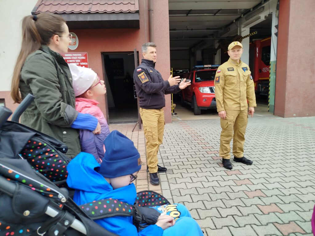 Dwoje strażaków w mundurach rozmawia z dziećmi przed remizą strażacką. W tle widać otwartą bramę garażu z czerwonymi pojazdami strażackimi.