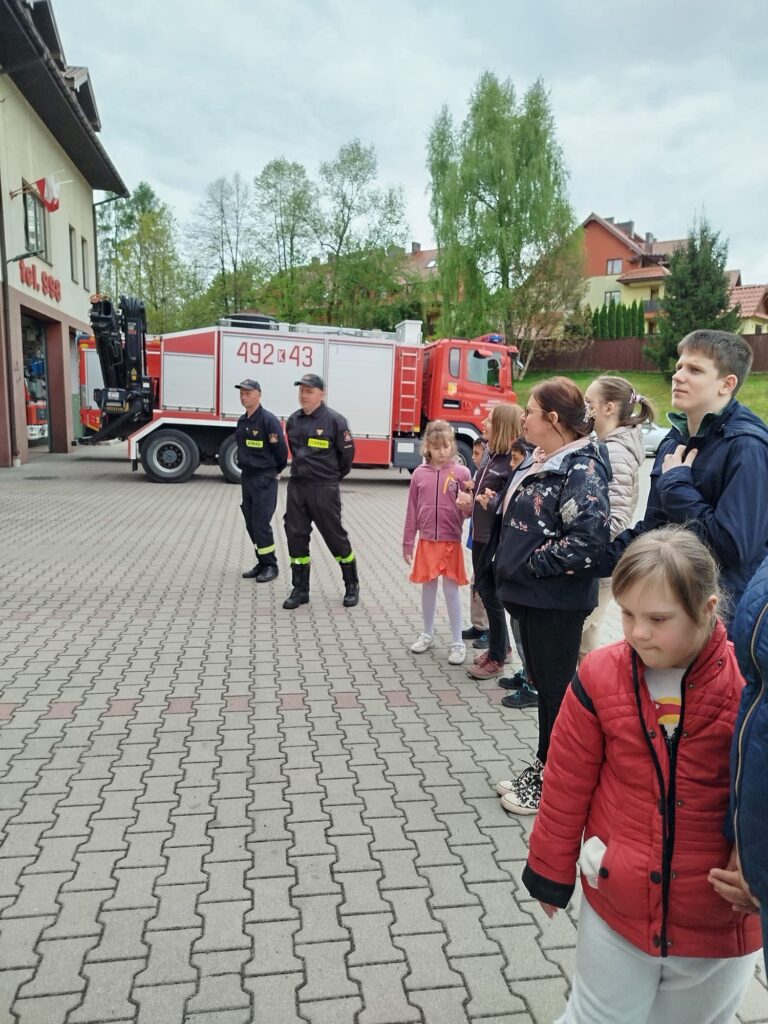 Grupa dzieci i dorosłych oraz dwóch strażaków stoi obok wozu strażackiego. Spotkanie odbywa się na placu przed remizą.
