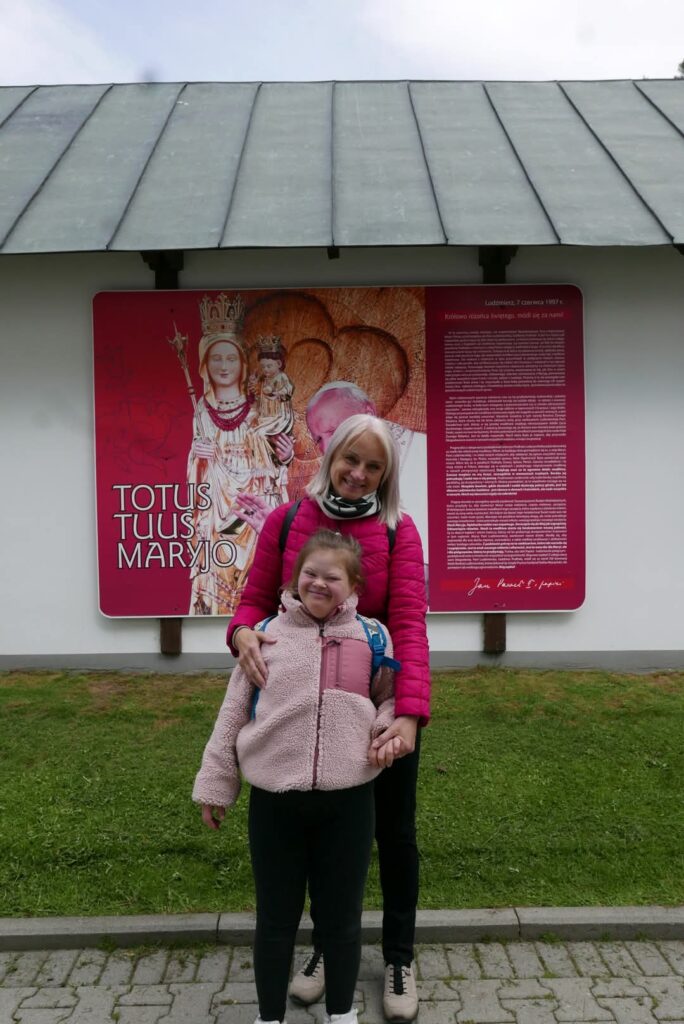 Uśmiechnięta kobieta i dziewczynka, stoją przed dużą tablicą z religijnym obrazem i napisem "Totus Tuus Maryjo".