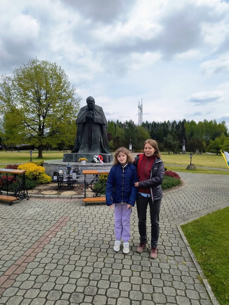 Nauczycielka z uczennicą stoją przed pomnikiem Jana Pawła II.