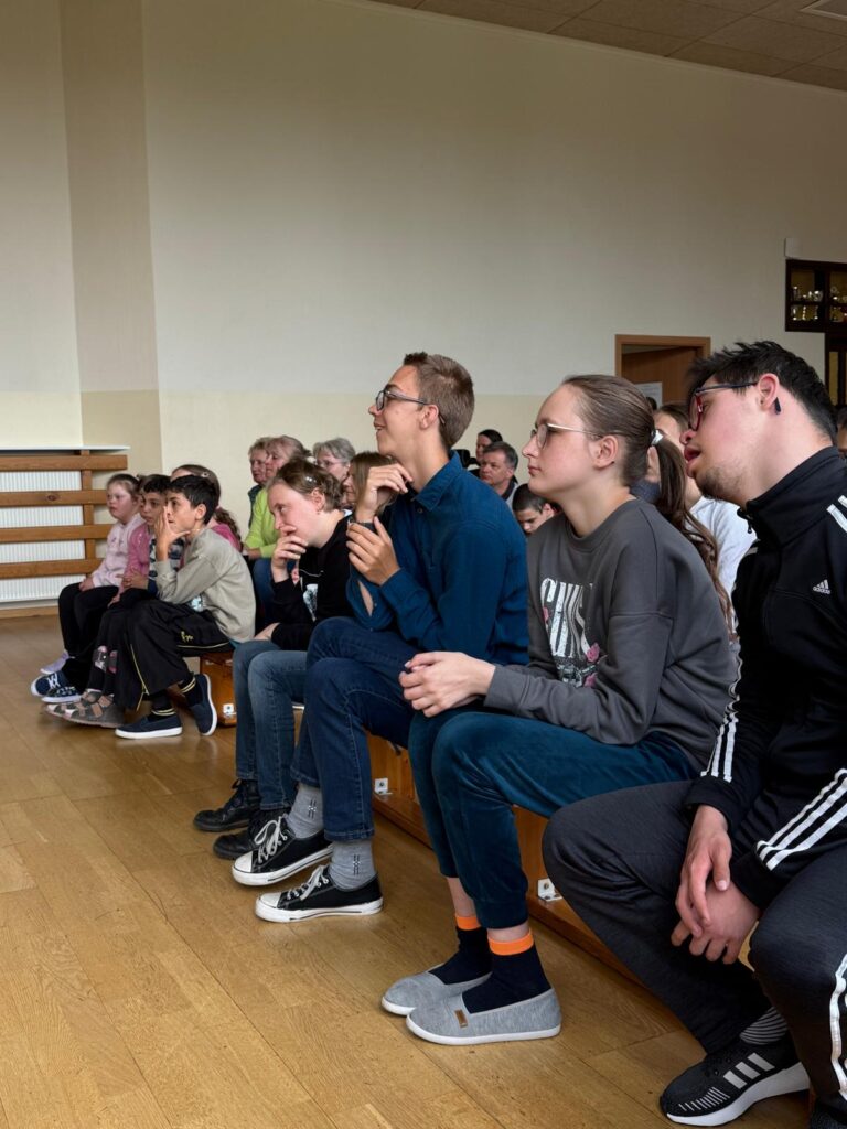 Duża grupa osób siedzi na ławkach z zainteresowaniem coś oglądając.