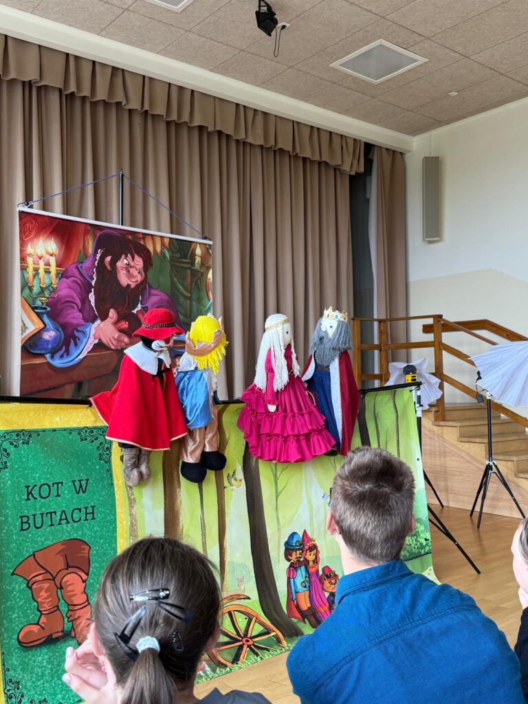 Duży plakat z postaciami z bajki nad którym widać kukiełki teatralne.