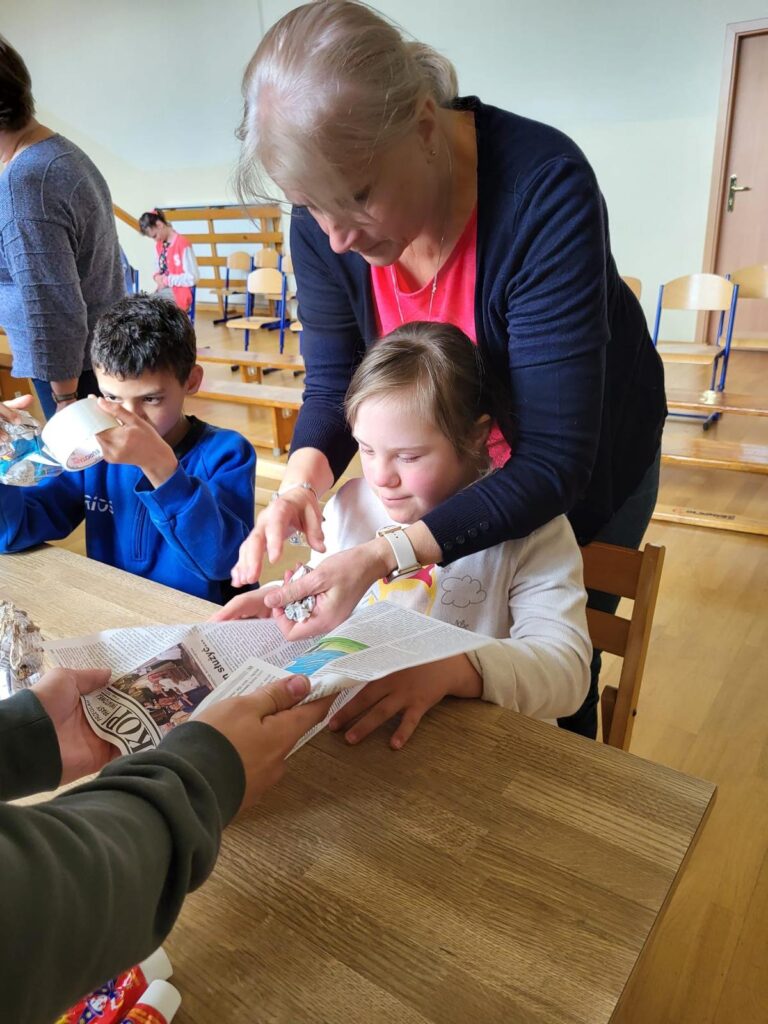 Dziewczynka i chłopiec siedzą przy stole, za dziewczynką stoi kobieta i pokazuje jej jak zrobić kulkę z gazety. Chłopiec okleja kulkę z gazety taśmą.