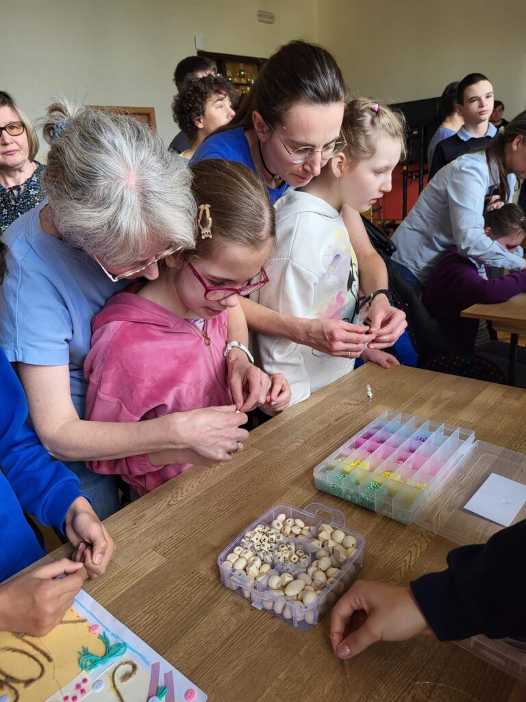Dwie dziewczynki stoją przy stole, za nimi stoją kobiety pomagając im nawlekać koraliki na żyłkę.