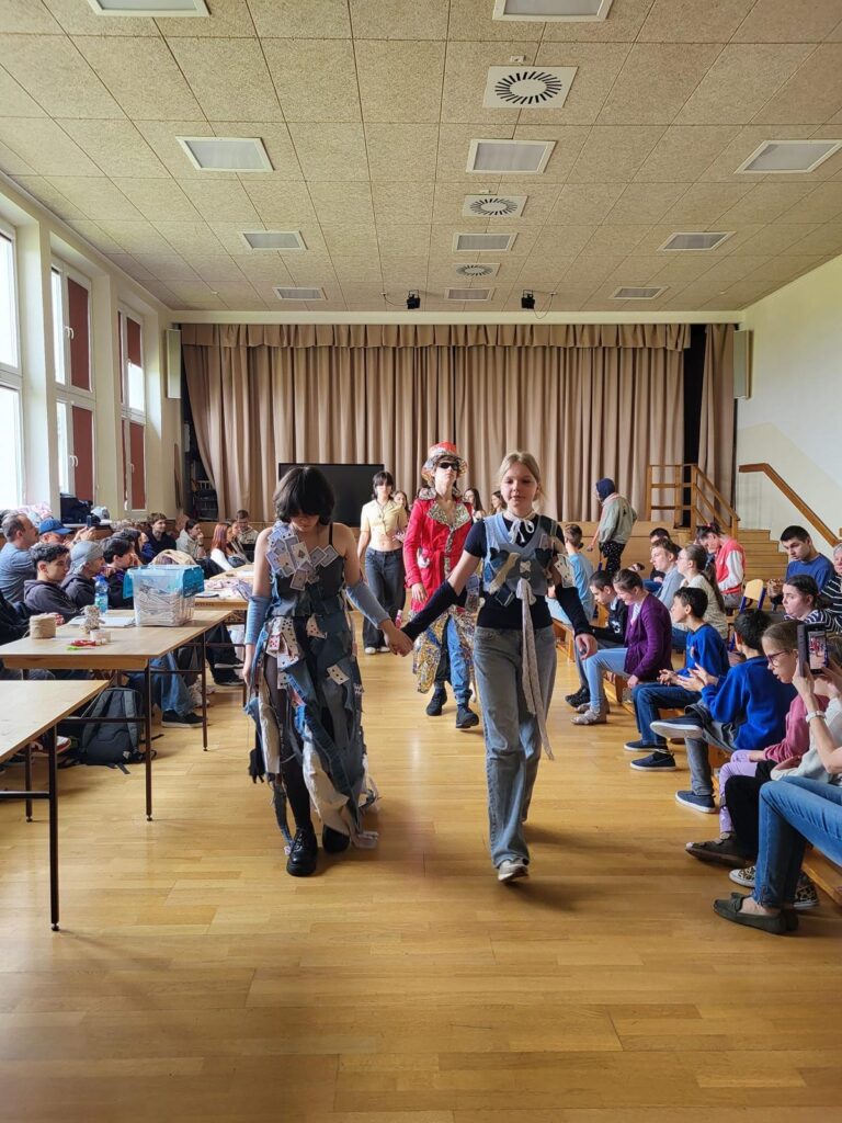 Grupa młodzieży w strojach z recyklingu jest na środku sali, po bokach grupa młodzieży i dorosłych obserwuje ich pokaz.