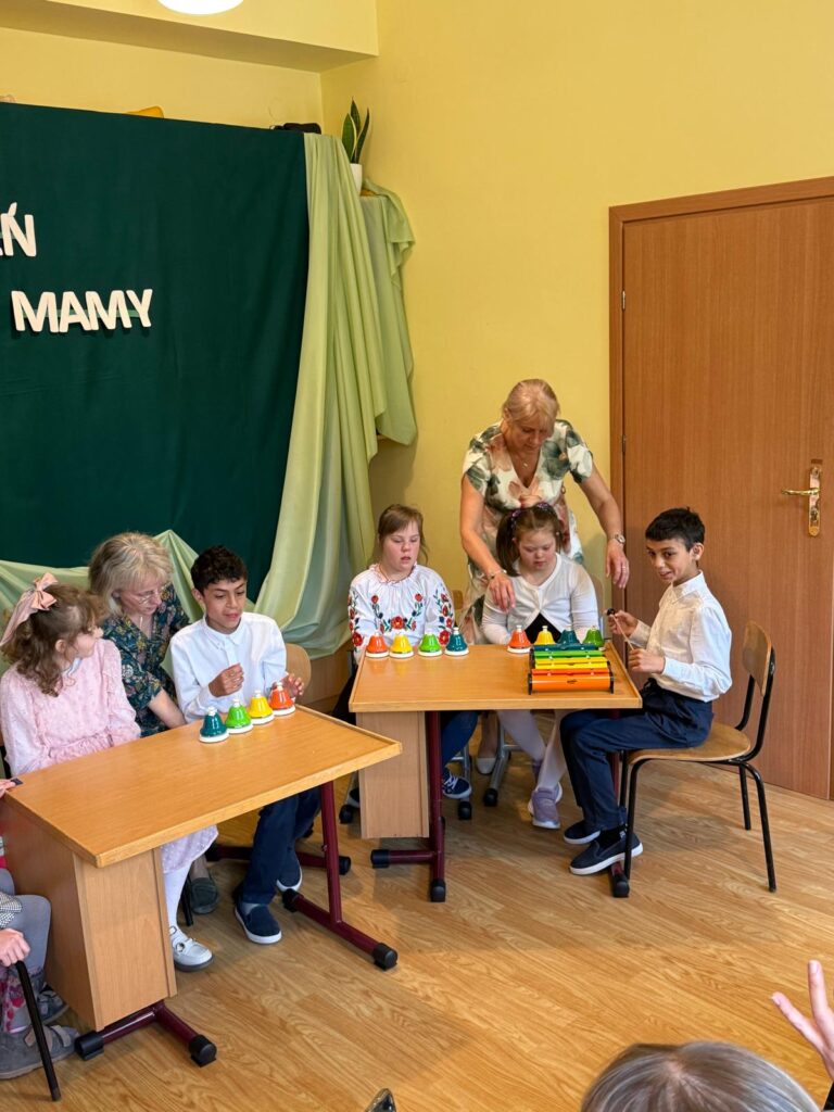 Grupa dzieci i dorosłych siedzi przy stolikach. Dzieci grają na kolorowych dzwonkach i cymbałkach, wspierane przez nauczycieli. W tle zielona dekoracja z napisem „DZIEŃ MAMY”.