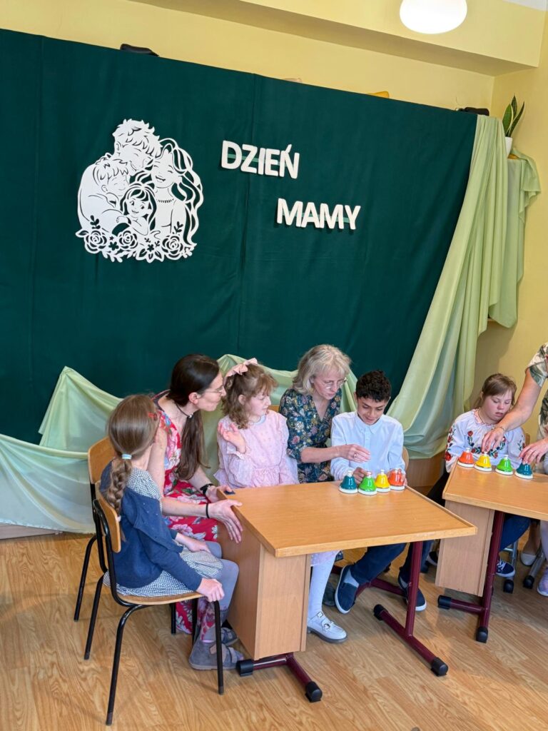 Dzieci i opiekunowie siedzą przy stolikach i grają na kolorowych dzwonkach. W tle widoczna dekoracja z napisem „DZIEŃ MAMY” oraz grafika przedstawiająca rodzinę.