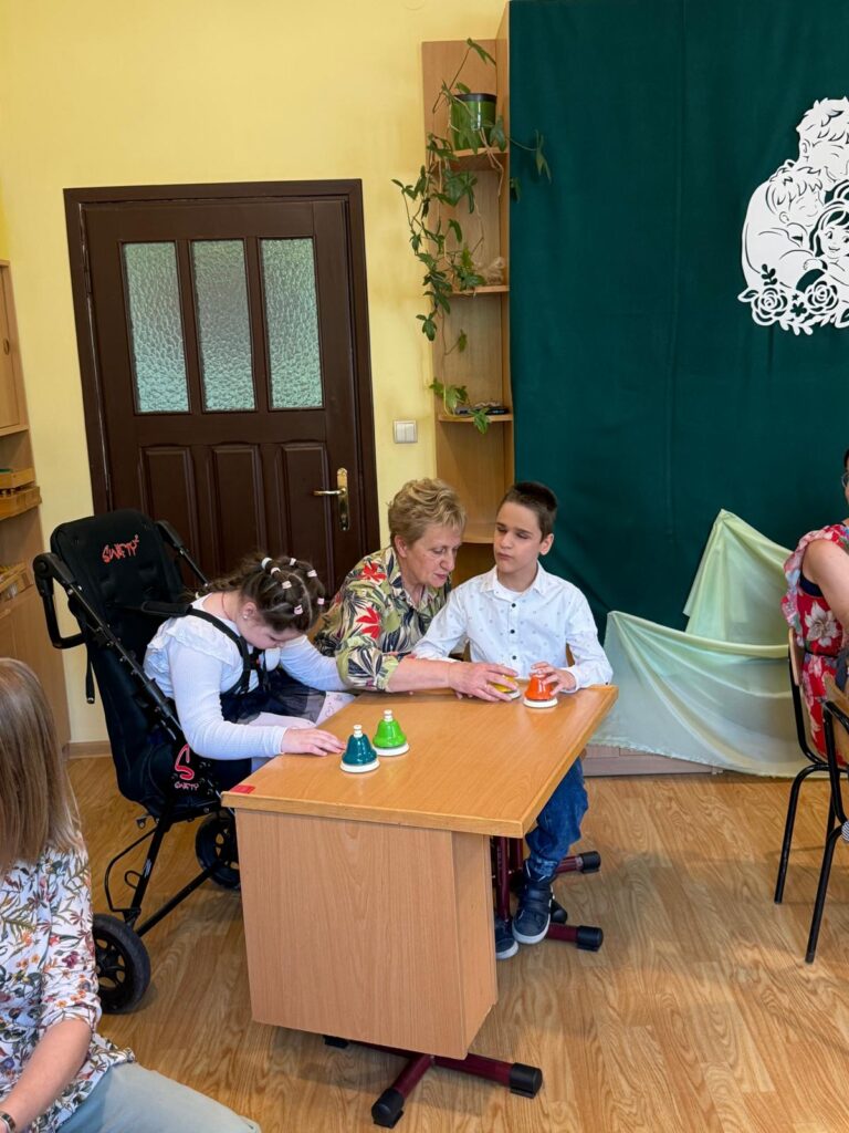 Kontynuacja sceny z występu na „Dzień Mamy” – dzieci i nauczyciel siedzą przy stoliku i grają na kolorowych dzwonkach. 