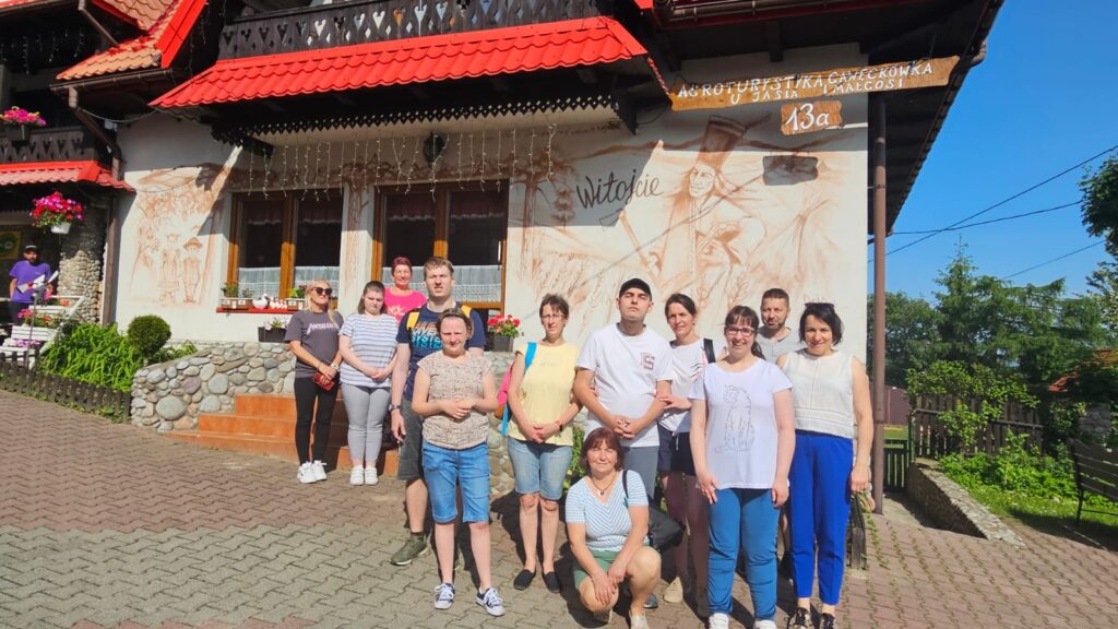 Grupa osób pozuje do wspólnego zdjęcia przed pensjonatem o tradycyjnej, góralskiej architekturze. Budynek ma czerwony dach, drewniany balkon i ozdobne malunki na ścianach. W tle widać napis „Agroturystyka u Jasia i Małgosi”.