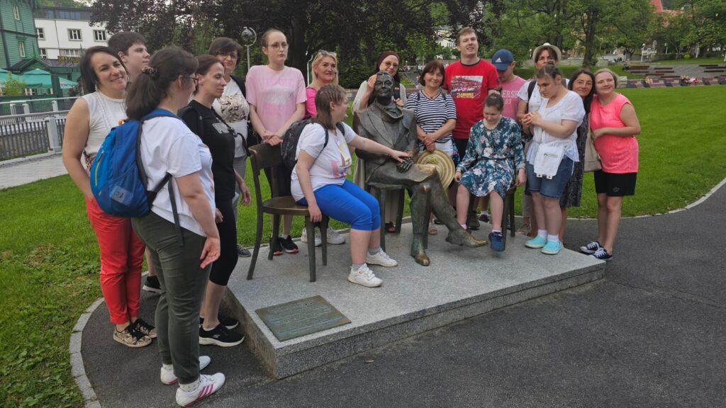 Grupa kilkunastu osób, w tym osoby z niepełnosprawnościami, zgromadzona wokół pomnika siedzącego mężczyzny. Uczestnicy wycieczki pozują do wspólnego zdjęcia w parku, w tle drzewa i zielony budynek.