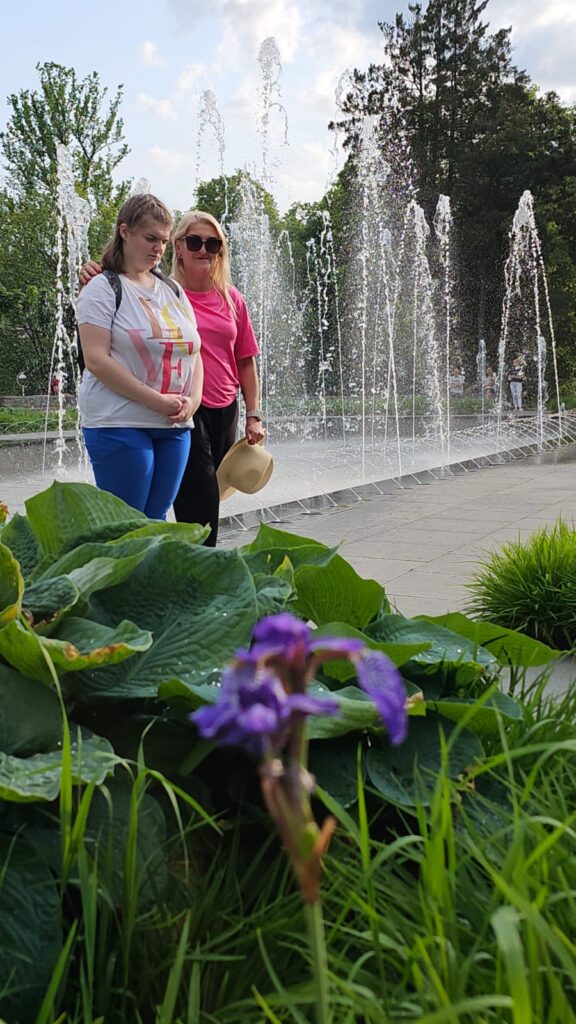 Dwie kobiety stoją przy fontannie w parku, otoczone zielenią i kwiatami; jedna trzyma kapelusz, a w tle widać tryskającą wodę i drzewa.